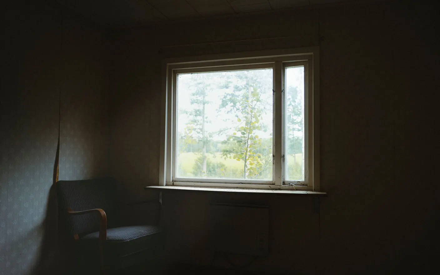 A chair beside a window in a dim room, looking out at a quiet outdoor scene.