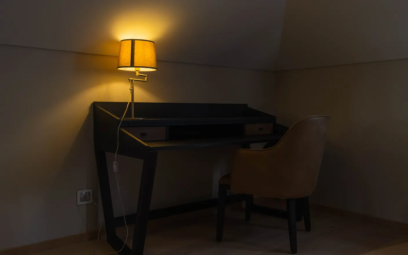 A dark room lit only by a warm desk lamp above a small writing desk and chair.