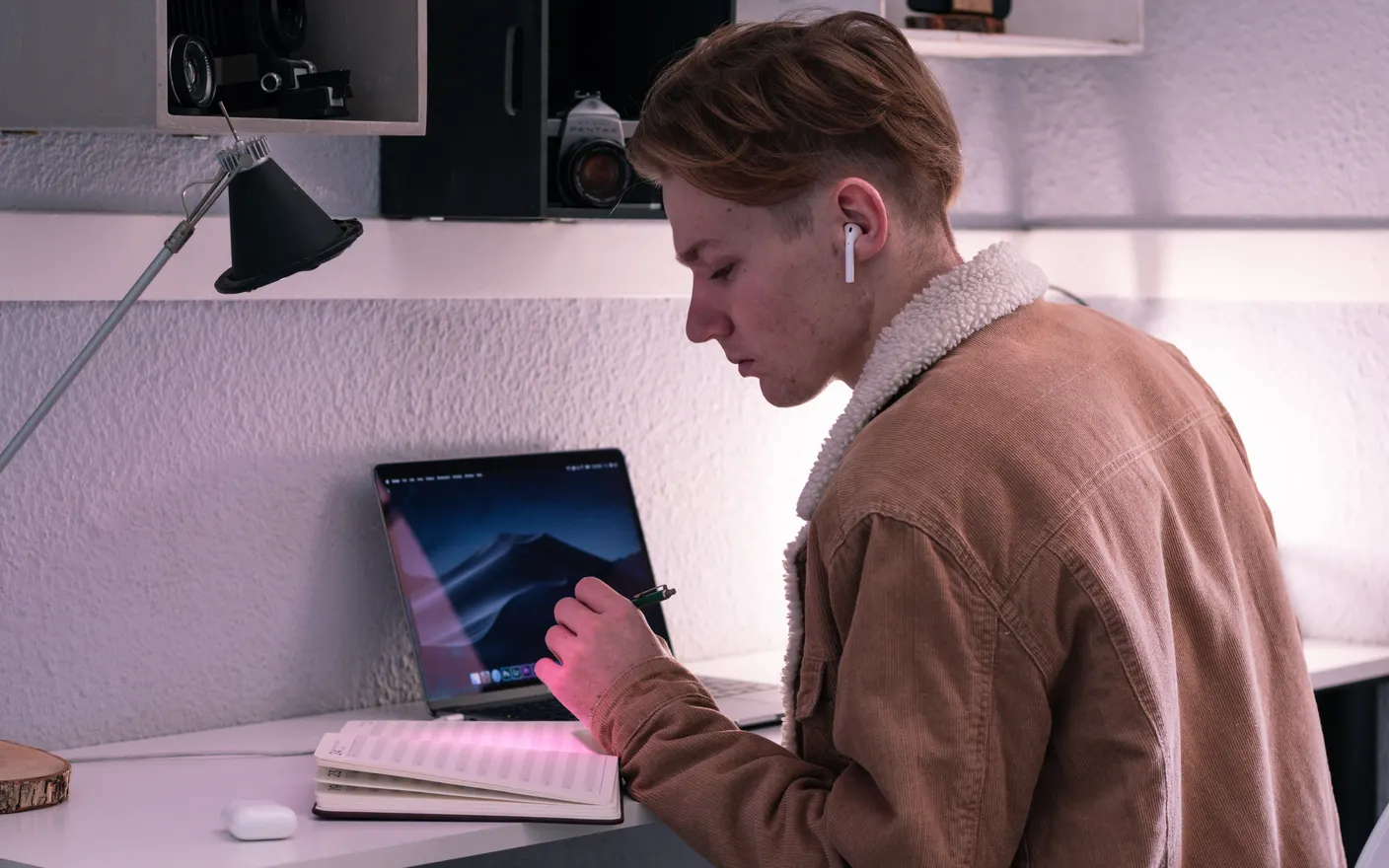 A student writing in a planner at a small desk beside a laptop, wearing earbuds.