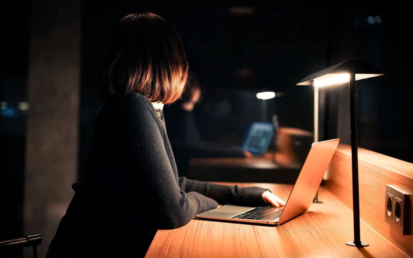 A woman working on a laptop at a desk at night, lit only by a warm lamp.