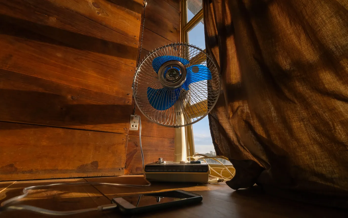 An electric fan standing beside a sunlit curtain in a warm room.