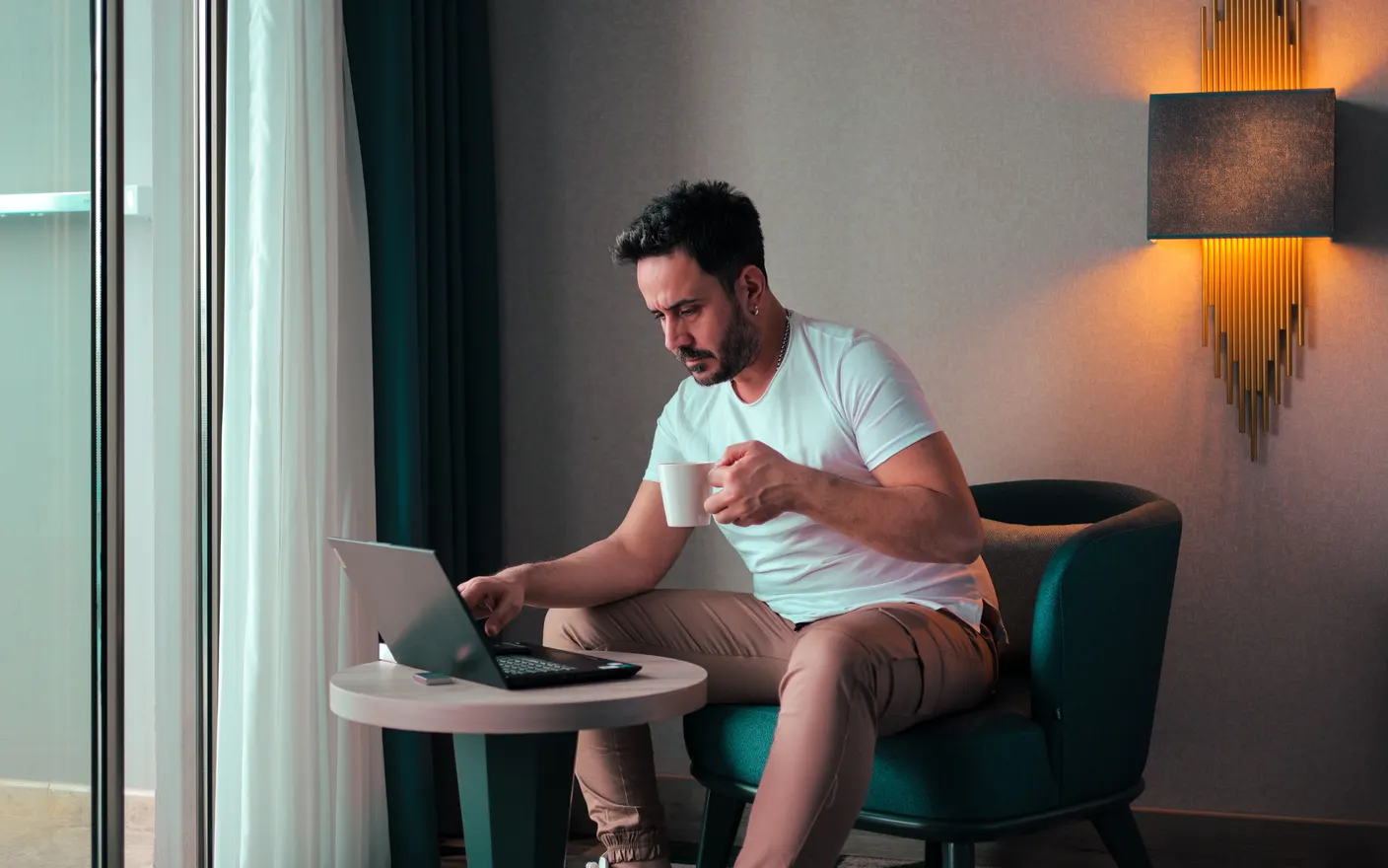 A person working on a laptop balanced on a small round side table in a hotel room, with coffee in hand.