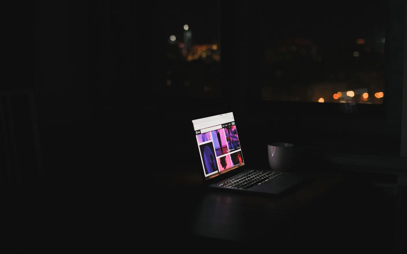 A laptop glowing on a desk in a dark room, with a city visible through the window behind it.