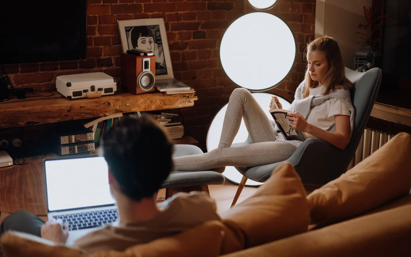Two people sharing a living room in the evening, one working on a laptop and one reading.
