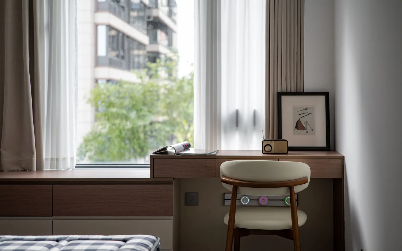 A narrow desk built along a bedroom window in a small apartment, with a chair, a radio and a framed print.