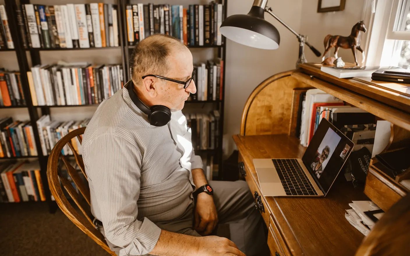 A man on a video call at a home desk with a bookshelf behind him.